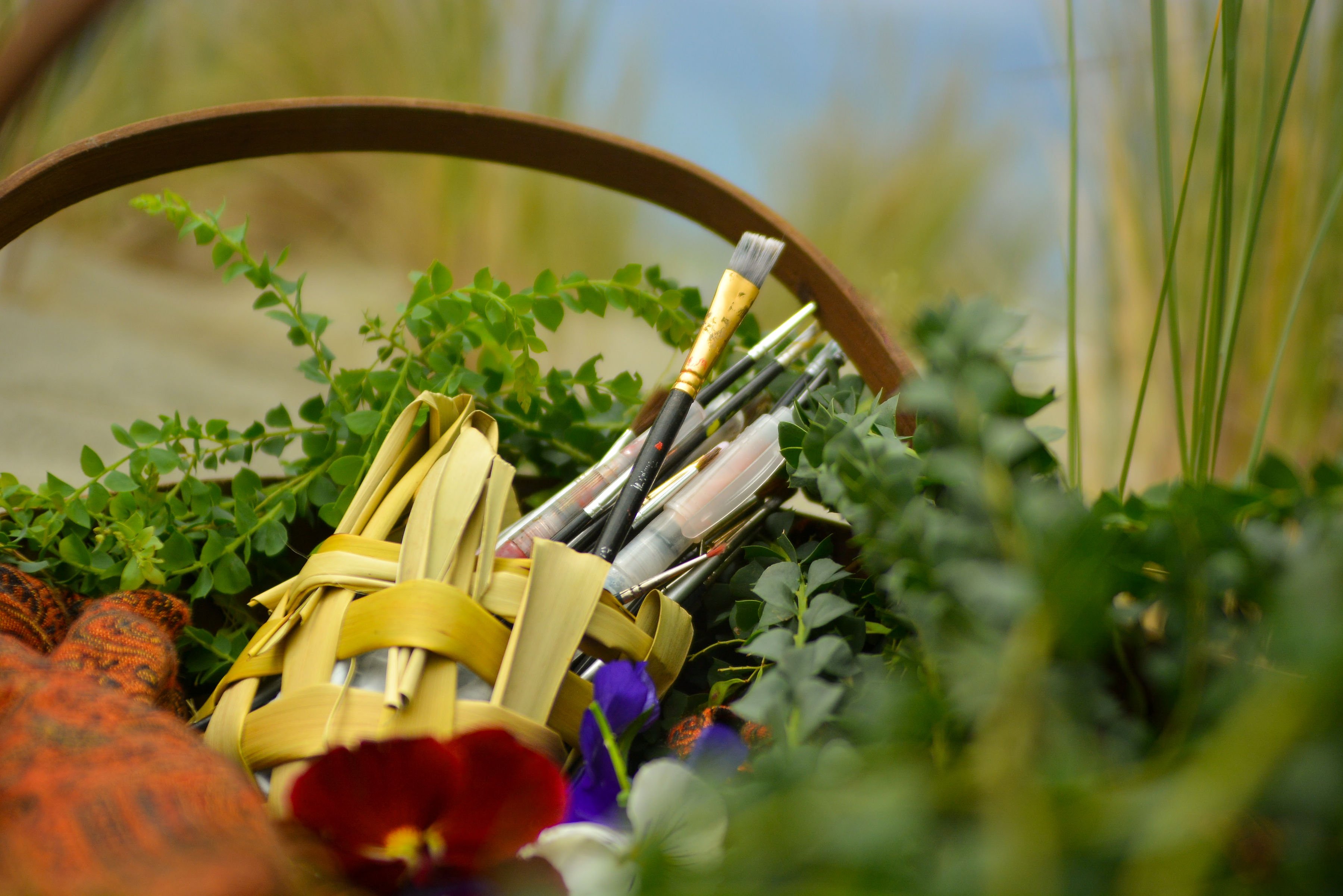A harakeke/flax weaved basket containing paintbrushes, surrounded by greenery.